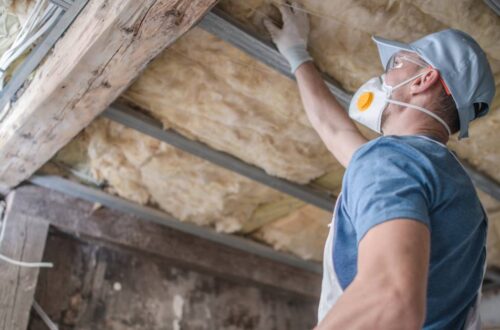 A man wearing protective gear like a respirator and gloves inspecting old, discolored insulation in a home.