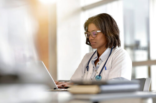 A doctor looking down at her laptop computer as she types, wearing a stethoscope and a white lab coat with a pen in the pocket.