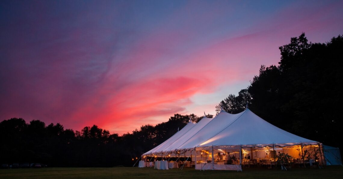 A vibrant orange, pink, and dark blue sunset over an event tent in a field. The tent has ambient lighting and tables.