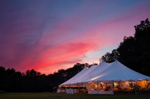 A vibrant orange, pink, and dark blue sunset over an event tent in a field. The tent has ambient lighting and tables.