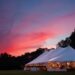 A vibrant orange, pink, and dark blue sunset over an event tent in a field. The tent has ambient lighting and tables.