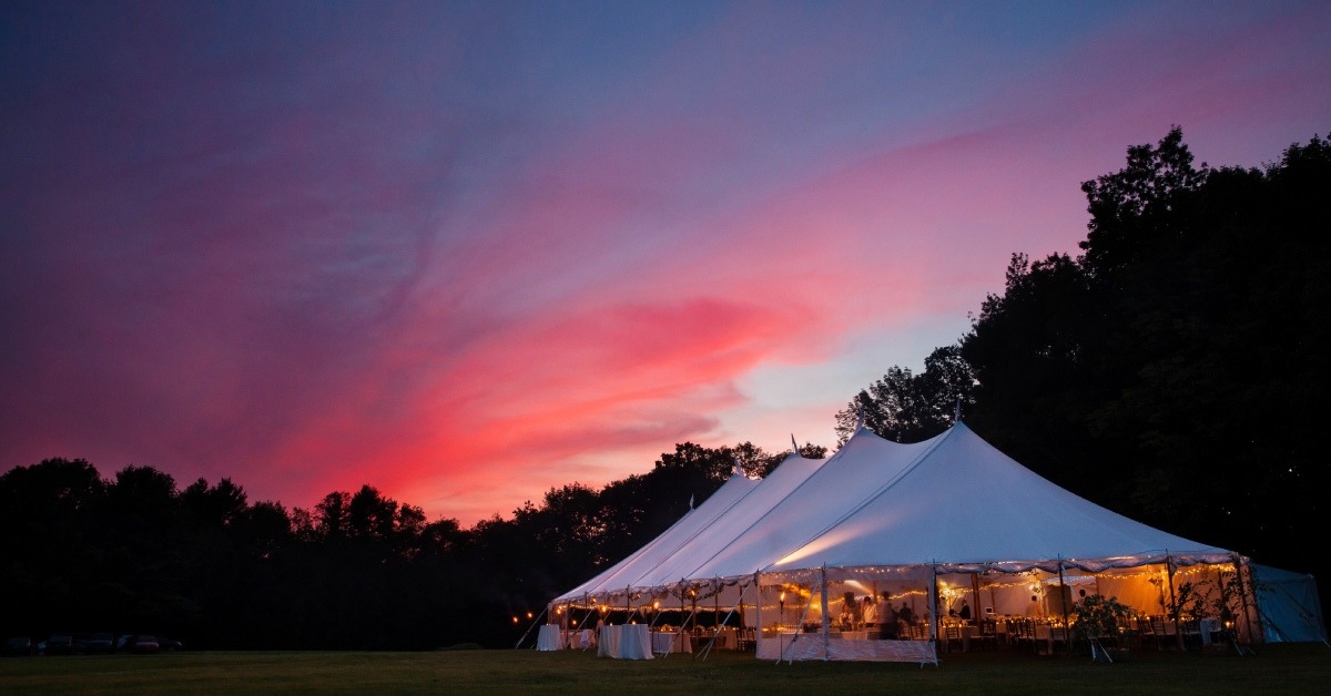 A vibrant orange, pink, and dark blue sunset over an event tent in a field. The tent has ambient lighting and tables.