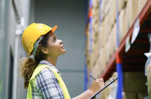 A worker does an inventory check on packages in a warehouse factory. She wears a yellow hard hat and jacket.