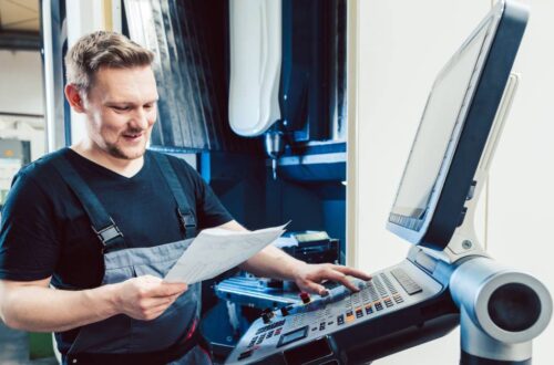 A worker wearing overalls in an industrial setting holding a piece of paper while entering instructions into a CNC machine.