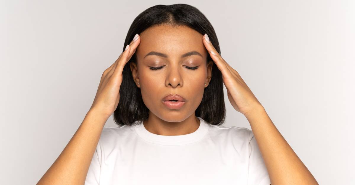 A woman wearing a white shirt in front of a white background holding her hands up to her forehead as she tries to destress.