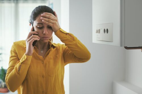 A woman on the phone who looks very stressed. She's standing next to her boiler and has her hand on her forehead.