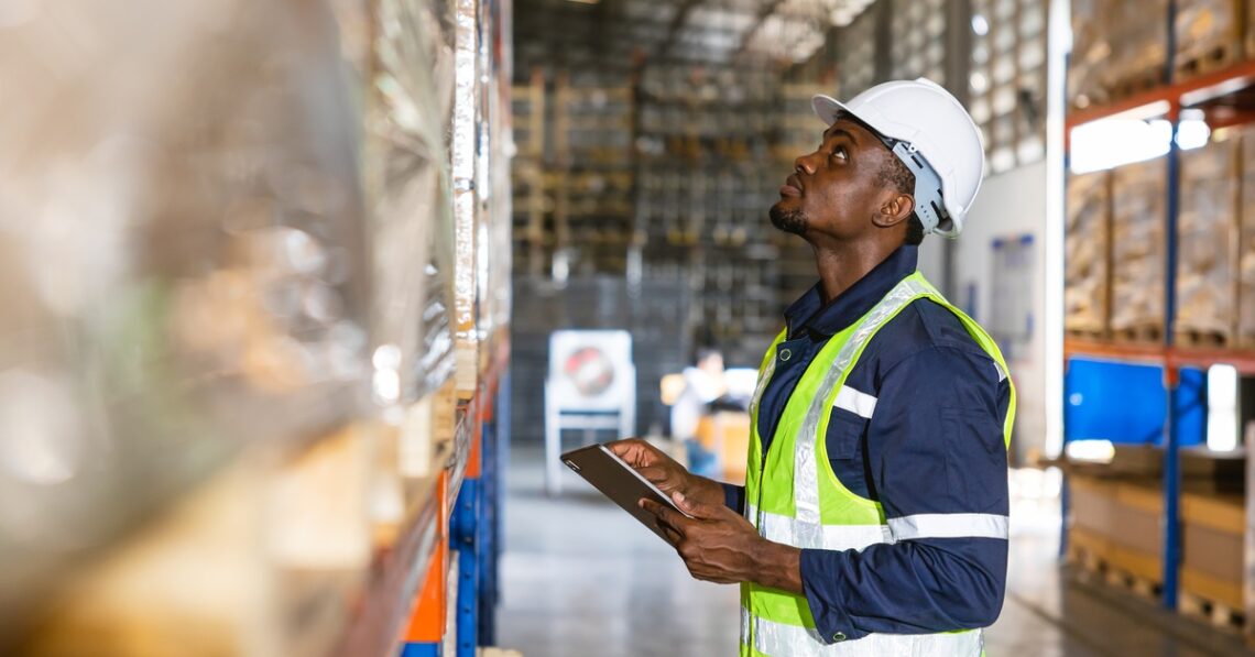 A warehouse worker wearing a hardhat and a reflective vest holding a clipboard. He is looking up at a shelf that's blurred.