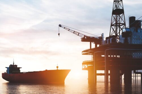 An oil platform sits in the ocean as a container ship approaches its general direction. The sun sets on the horizon.