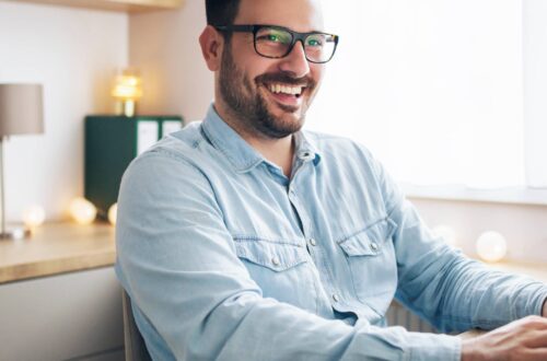 A remote worker in his home office types on a black keyboard while looking at his monitor and smiling.