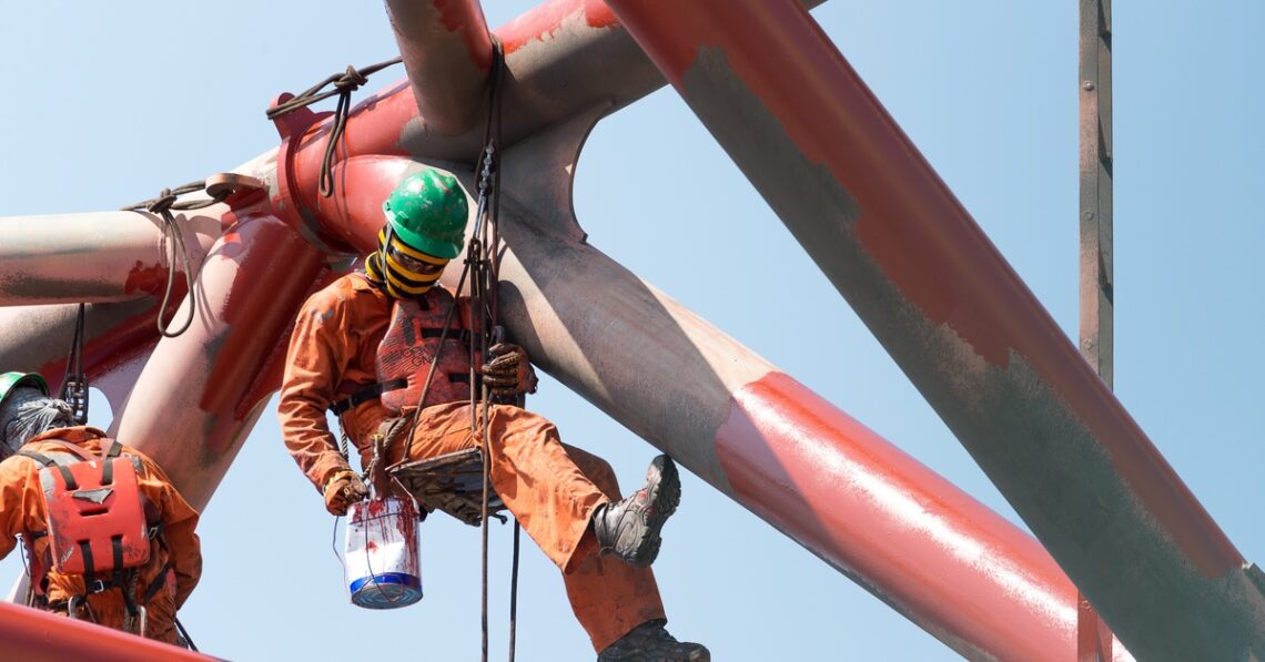 Two workers wearing orange jumpsuits and green protective hats paint a structure while being held up by a cable rig.