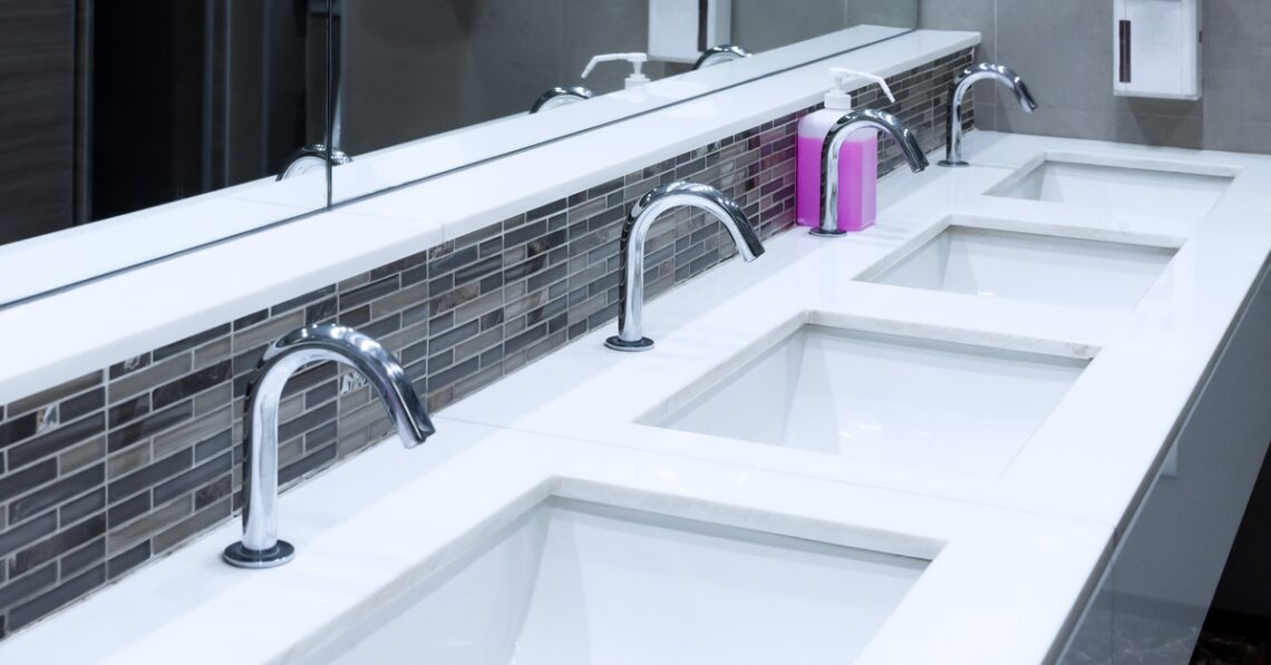 A line of white sinks inside a commercial restroom has silver automatic faucets and an oversized mirror.