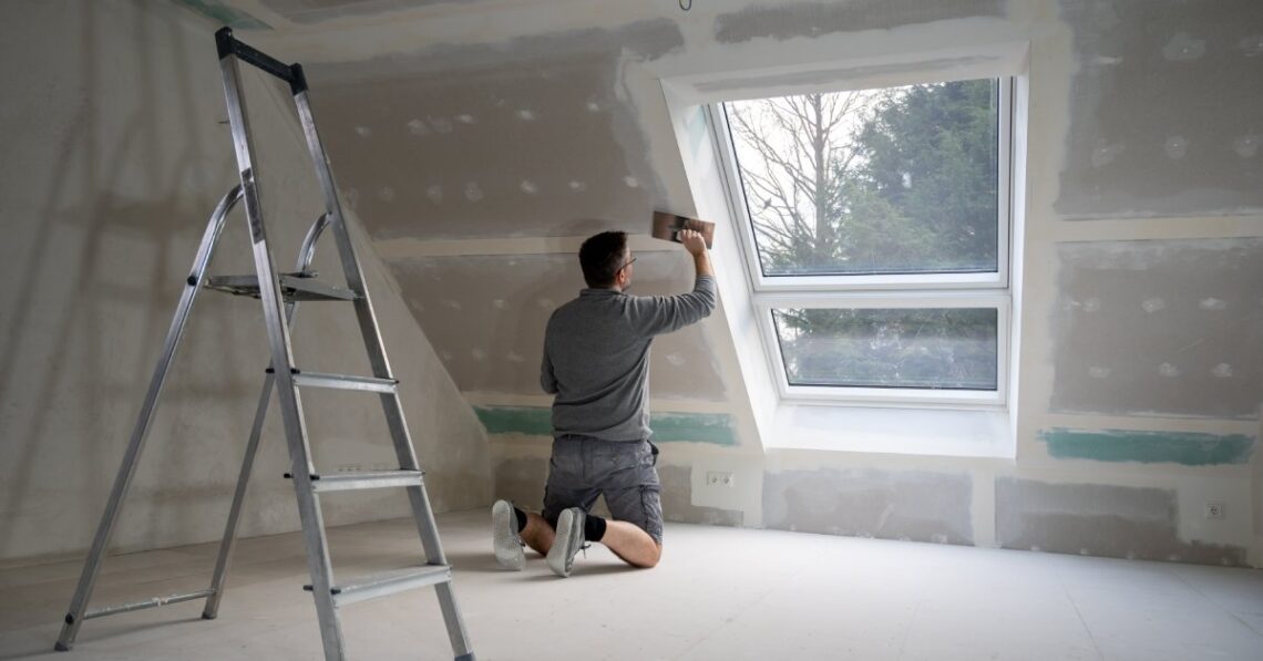A man wearing a gray shirt and gray shorts uses a scraper to apply a white paste onto a bare home wall beside a window.