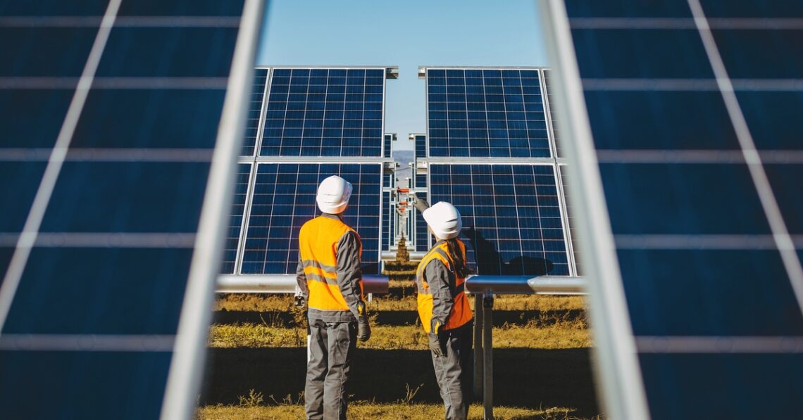 Two workers standing outside are examining solar panels arranged in a group with rows extending far back.