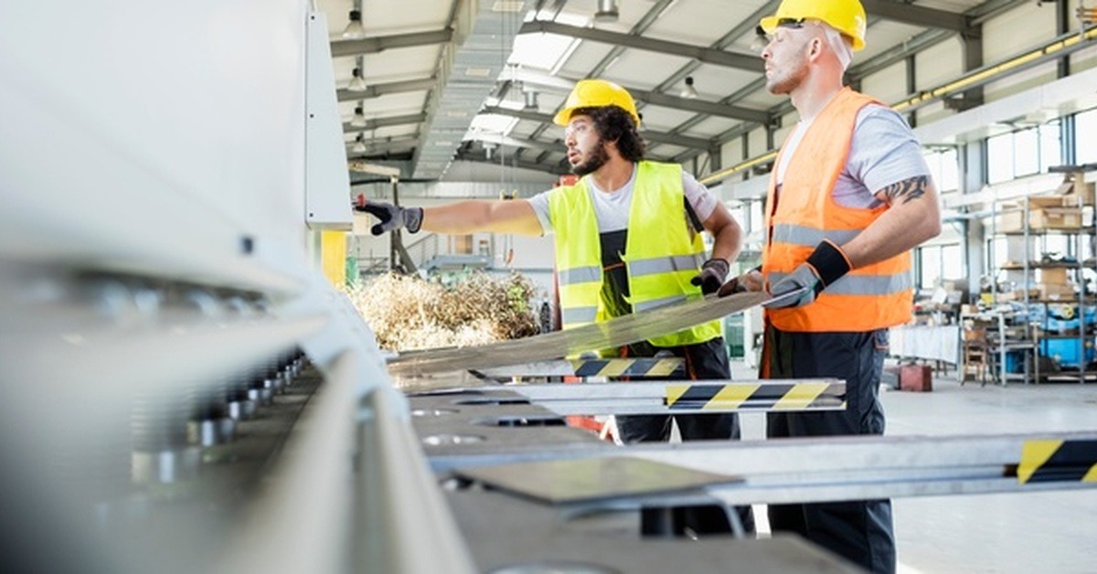 Two men in a manufacturing setting wearing high-visibility vests yellow hard hats while working with machinery.