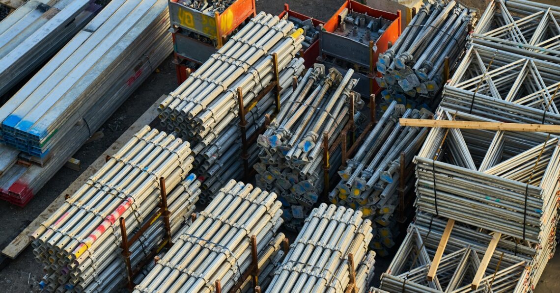 Stacks of steel, iron, and aluminum bars and frames in varying heights sit on dirt, lit by daylight.