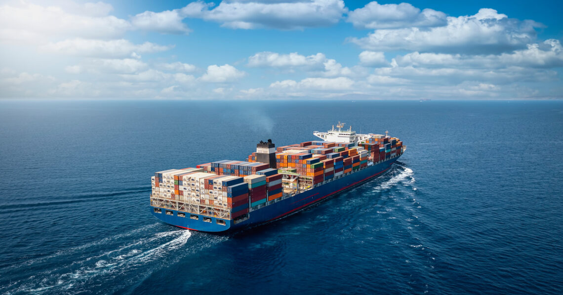 A large container cargo ship travels over a blue ocean. There are numerous colorful shipping containers on top of the ship.