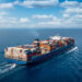 A large container cargo ship travels over a blue ocean. There are numerous colorful shipping containers on top of the ship.