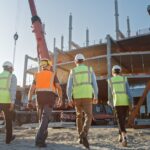 Four workers in safety vests and hard hats walking across a construction site toward an unfinished building. Construction Company Can Save Money