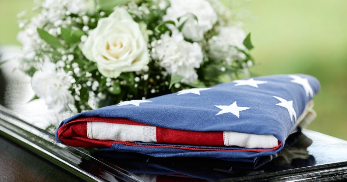 A US flag is folded neatly into a triangle on top of a black coffin next to a bouquet of white roses.