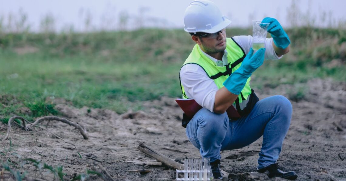 A man in safety gear squats near a water source, holding a clear cup of green liquid beside a rack of test tubes.