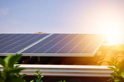 Tree branches frame a red roof as sunlight peeks over photovoltaic panels beside white gutters and trim.