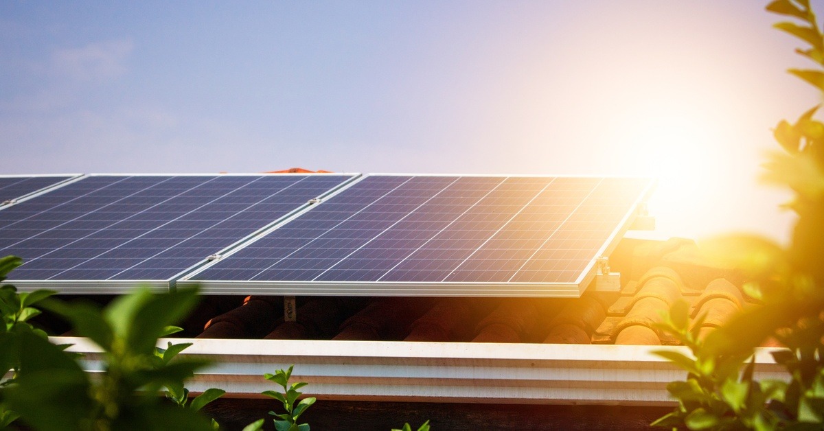 Tree branches frame a red roof as sunlight peeks over photovoltaic panels beside white gutters and trim.