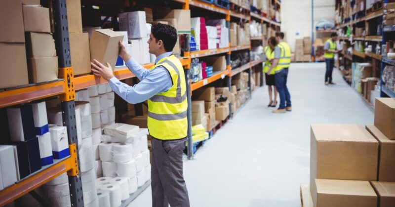 Warehouse operators inspecting boxes of products on shelves. There is a pallet of boxes in the middle of the aisle.
