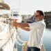 An older man is securing his sailboat at a marina, pulling the rope tight to prevent the boat from drifting.