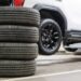 A white SUV is lifted up in a shop, and its trunk is open. A used tire stack sits on the ground next to it.