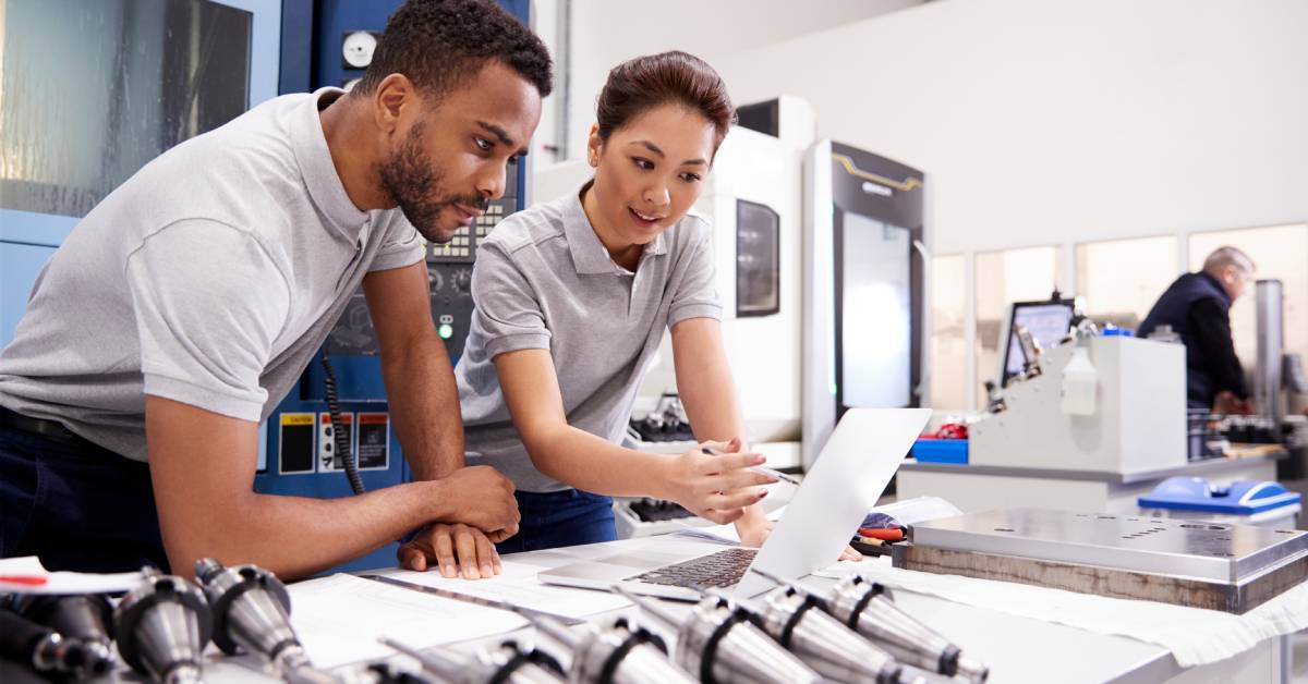 Two engineers, one male and female, in a laboratory examine a laptop with metal components on the desk in front of them.