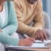 A senior man and woman sit next to each other on a couch while looking over documents on a coffee table.