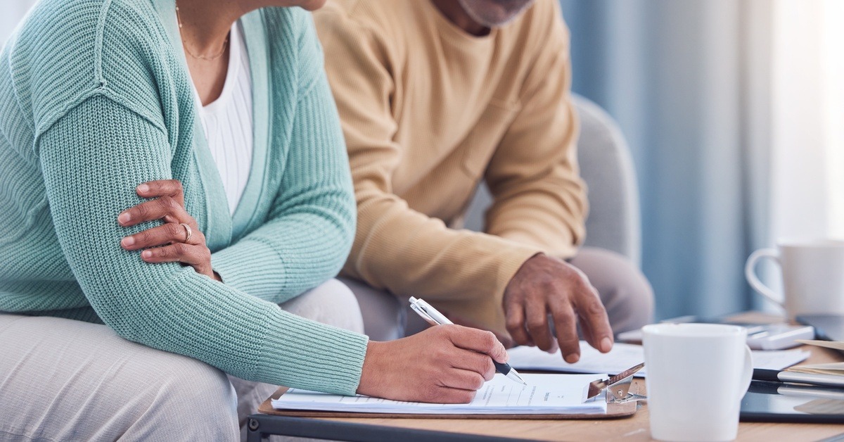 A senior man and woman sit next to each other on a couch while looking over documents on a coffee table.