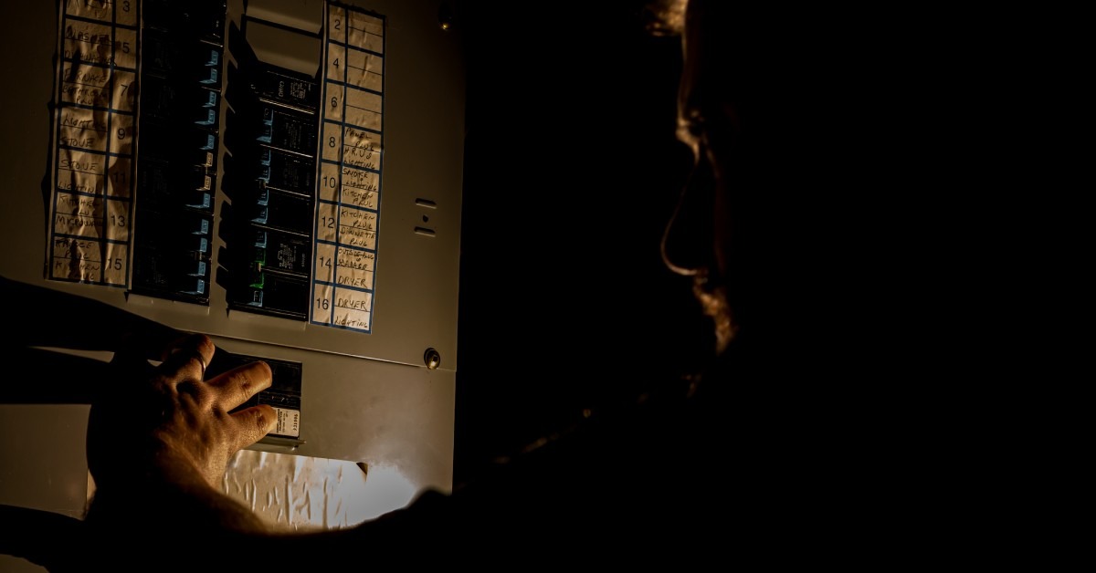 A person stands in the dark next to a dimly lit fuse box with fourteen black and blue switches visible.