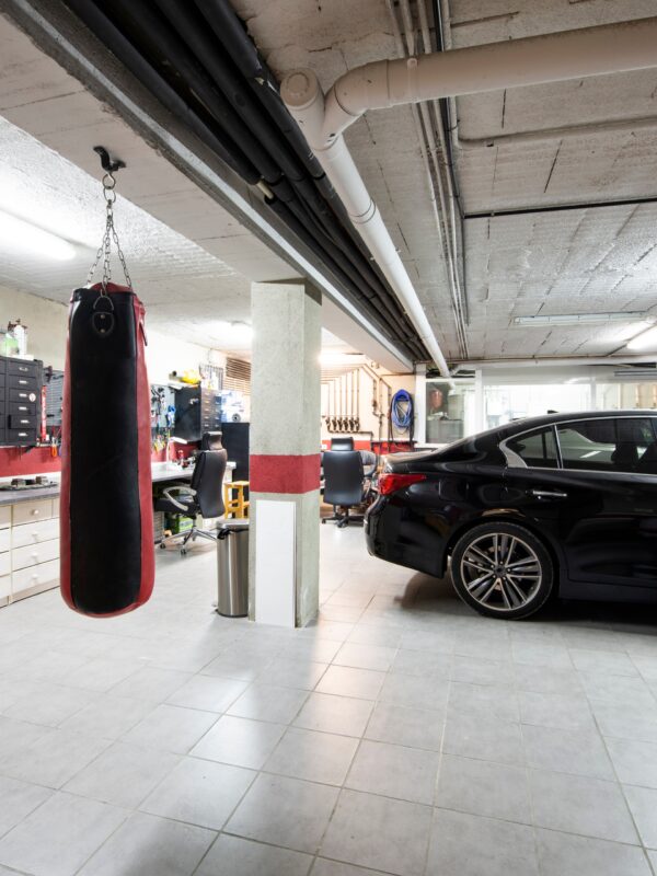 A black sedan and a white car are parked inside a garage with cabinetry, a punching bag, and tile flooring visible.