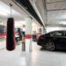 A black sedan and a white car are parked inside a garage with cabinetry, a punching bag, and tile flooring visible.