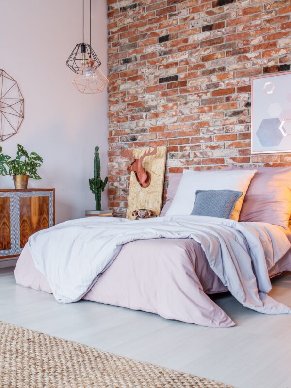 An industrial bedroom with an exposed brick wall and metal hanging pendant lights. There's a wooden chest left of the bed.