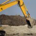 A person operates a yellow excavator on a site surrounded by dirt, while a man in an orange vest stands in front, watching.