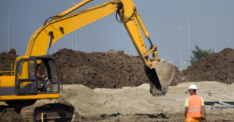 A person operates a yellow excavator on a site surrounded by dirt, while a man in an orange vest stands in front, watching.