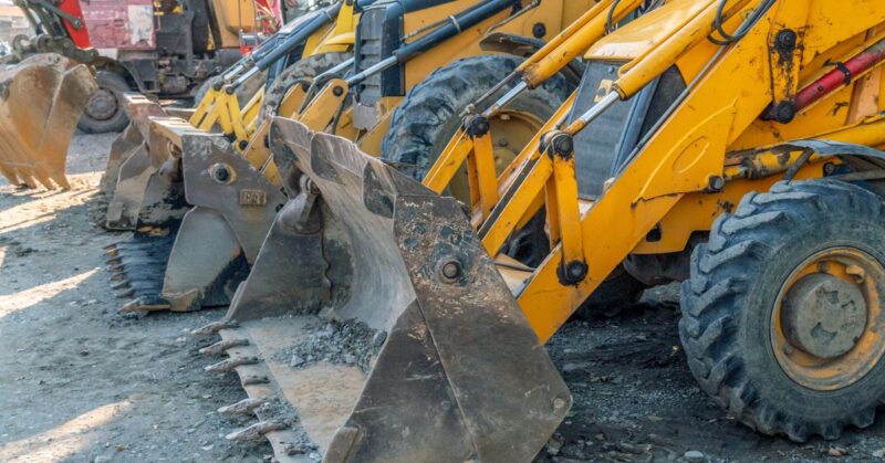 Yellow bulldozers sit in a row with dirt-covered tires and buckets resting on the ground filled with debris.