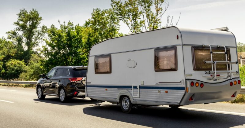 A caravan trailer on a freeway road. A Black SUV is carrying the caravan on the highway near a ramp.