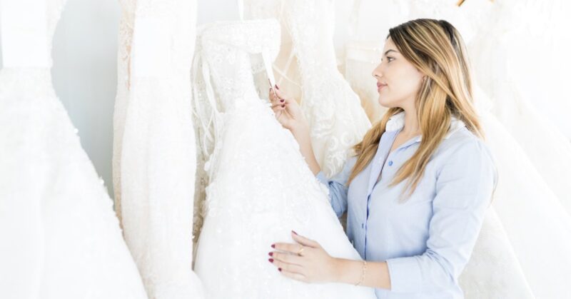 A woman checking the price tag on a wedding dress hanging from a rack among other gowns in a bright white room.