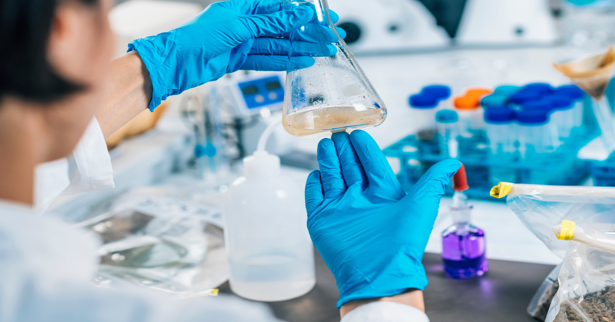 A close-up of a scientist wearing a white coat and blue gloves testing a substance in a beaker and swishing it around.