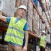A warehouse employee is pointing at a barcode on a shelf holding pallets. He is looking at another employee holding a tablet.