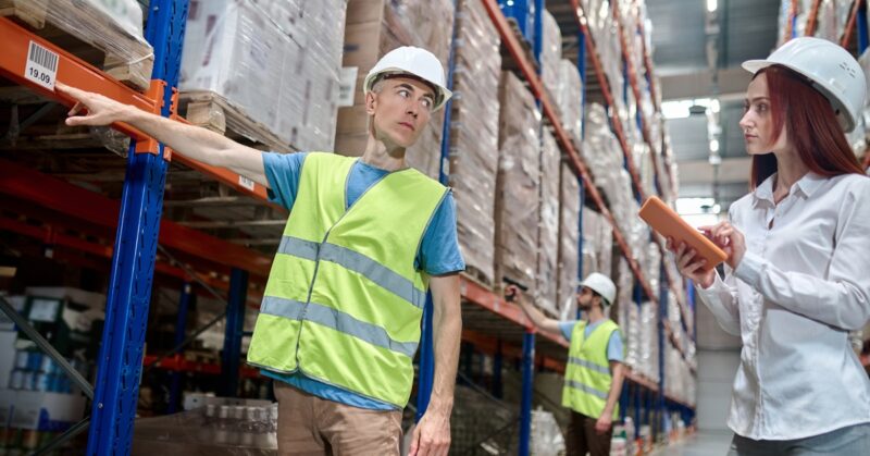 A warehouse employee is pointing at a barcode on a shelf holding pallets. He is looking at another employee holding a tablet.