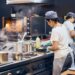 Kitchen staff wearing white t-shirts and fabric hairnets working inside a large kitchen with stainless steel appliances.