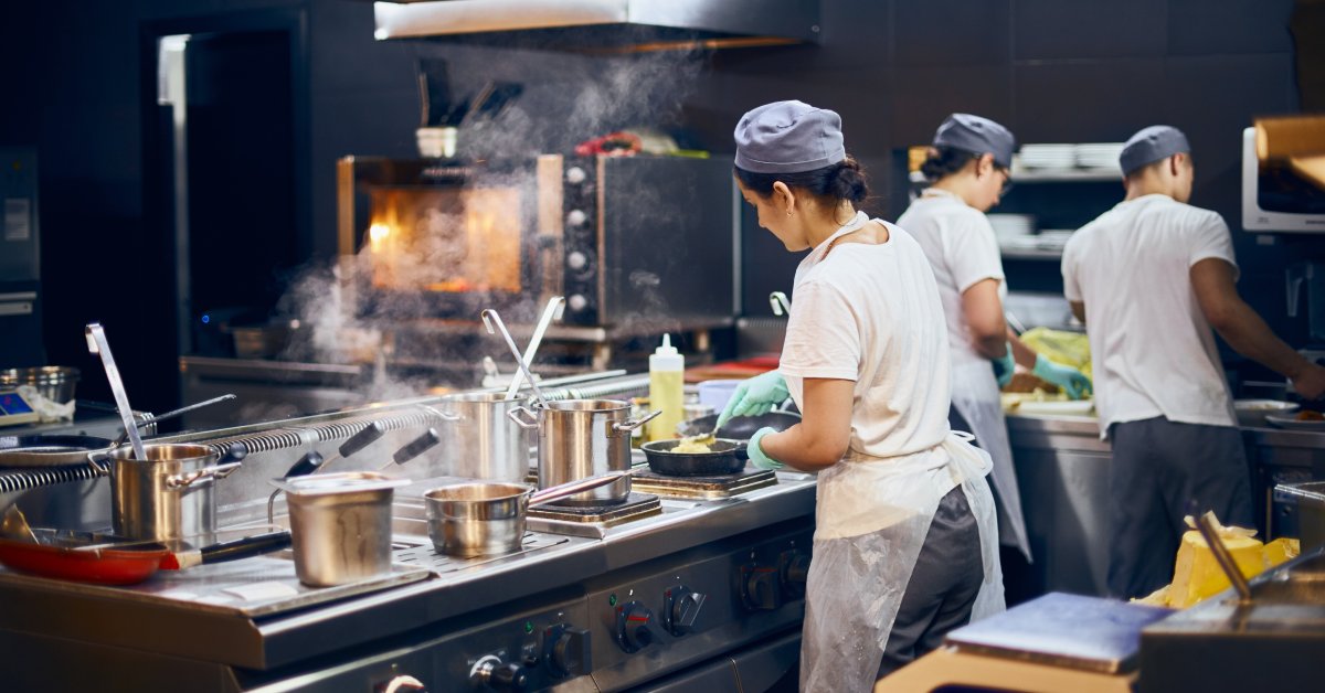 Kitchen staff wearing white t-shirts and fabric hairnets working inside a large kitchen with stainless steel appliances.