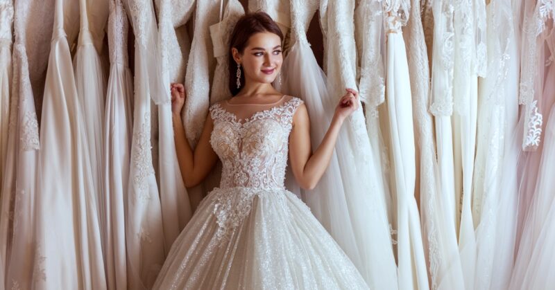 A woman in a wedding dress with a lace bodice and A-line skirt leans against a rack filled with countless wedding gowns.