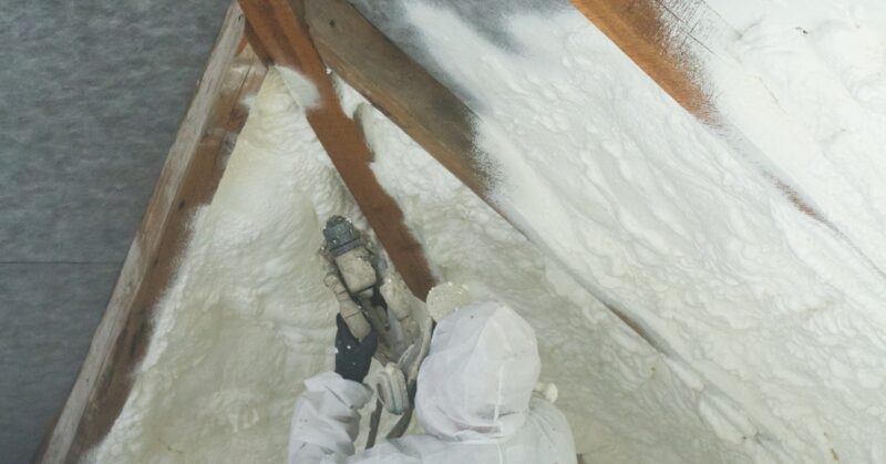 A contractor wearing a white protective suit applies spray foam insulation to the ceiling of an attic.