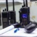 A gray desk holds an open book with a pen on top and several two-way radios clustered together on chargers.