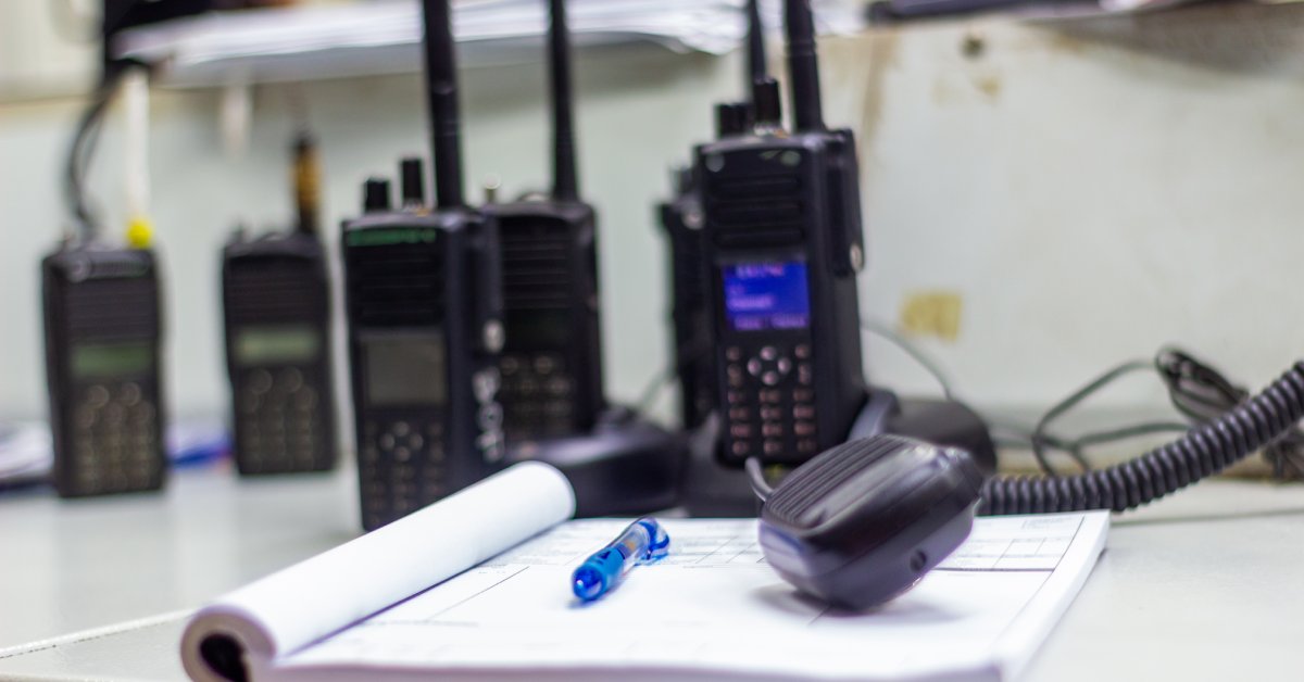 A gray desk holds an open book with a pen on top and several two-way radios clustered together on chargers.